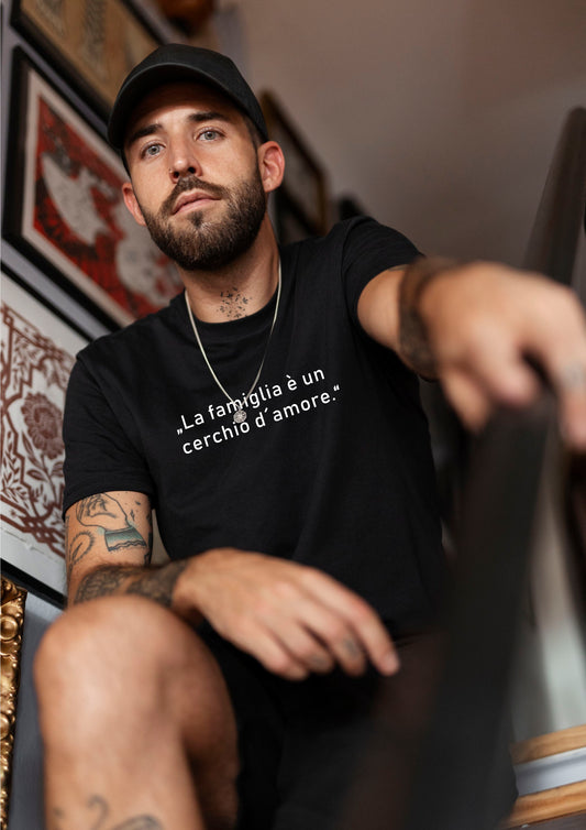 Person mit Tattoos und schwarzer Cap sitzt auf einer Treppe und trägt schwarzes T-Shirt mit weißem italienischen Zitat „La famiglia è un cerchio d’amore“