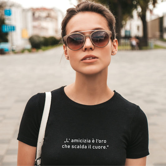 Person mit Sonnenbrille trägt schwarzes T-Shirt mit weißem italienischen Zitat „L’amicizia è l’oro che scalda il cuore“, fotografiert auf einer Straße mit Gebäuden und Bäumen im Hintergrund
