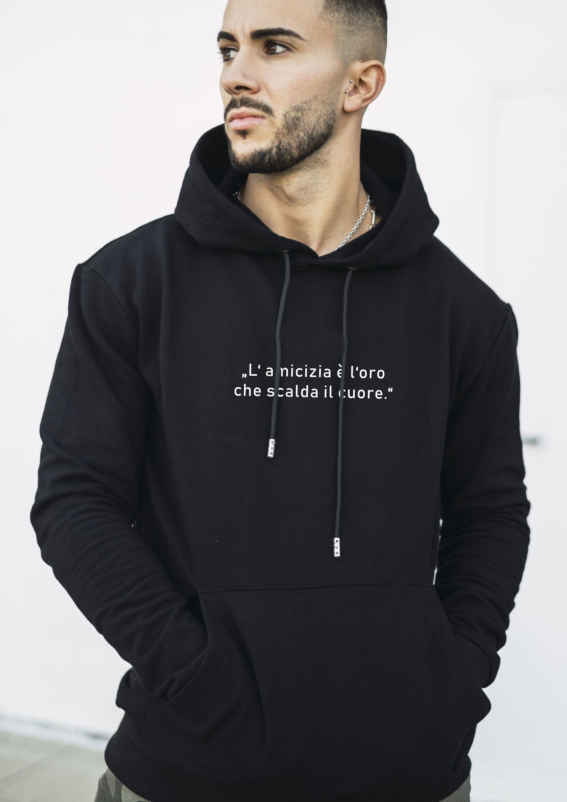 Person trägt schwarzen Hoodie mit italienischem Zitat „L’amicizia è l’oro che scalda il cuore“ auf der Vorderseite, Hände in der Kängurutasche