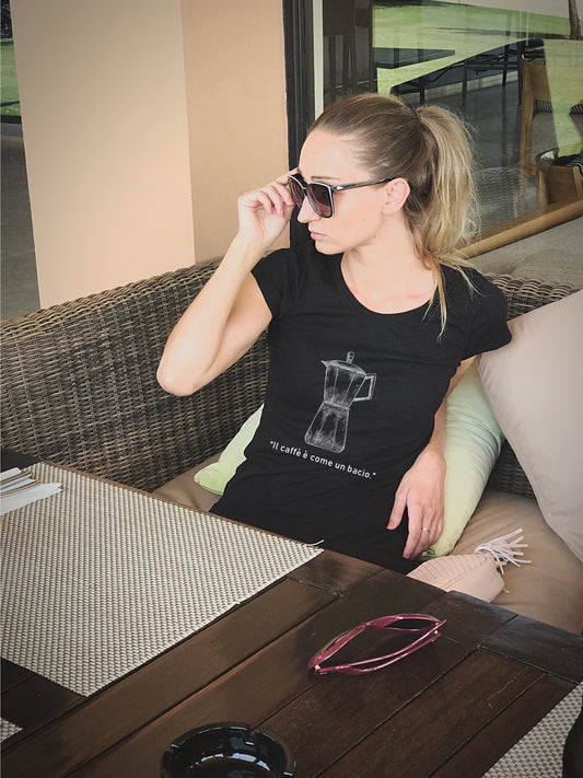Frau sitzt entspannt in Café-Atmosphäre und trägt schwarzes T-Shirt mit weißem Moka-Print und italienischem Spruch „Il caffè è come un bacio“