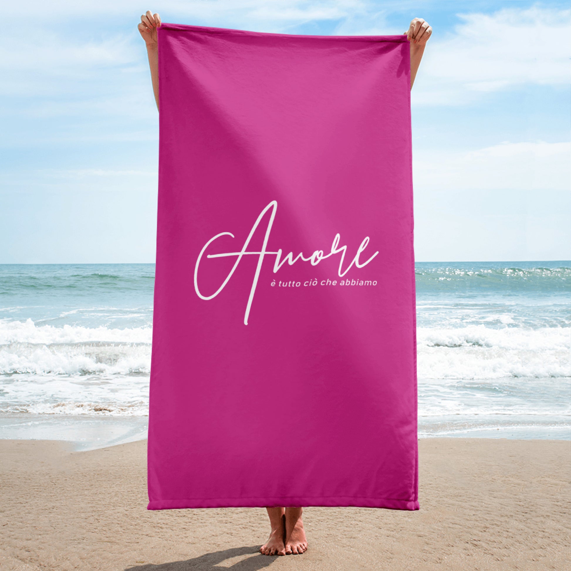 Person hält pinkes Handtuch mit weißem Schriftzug „Amore è tutto ciò che abbiamo“ am Strand – Liebesbotschaft trifft italienisches Design.