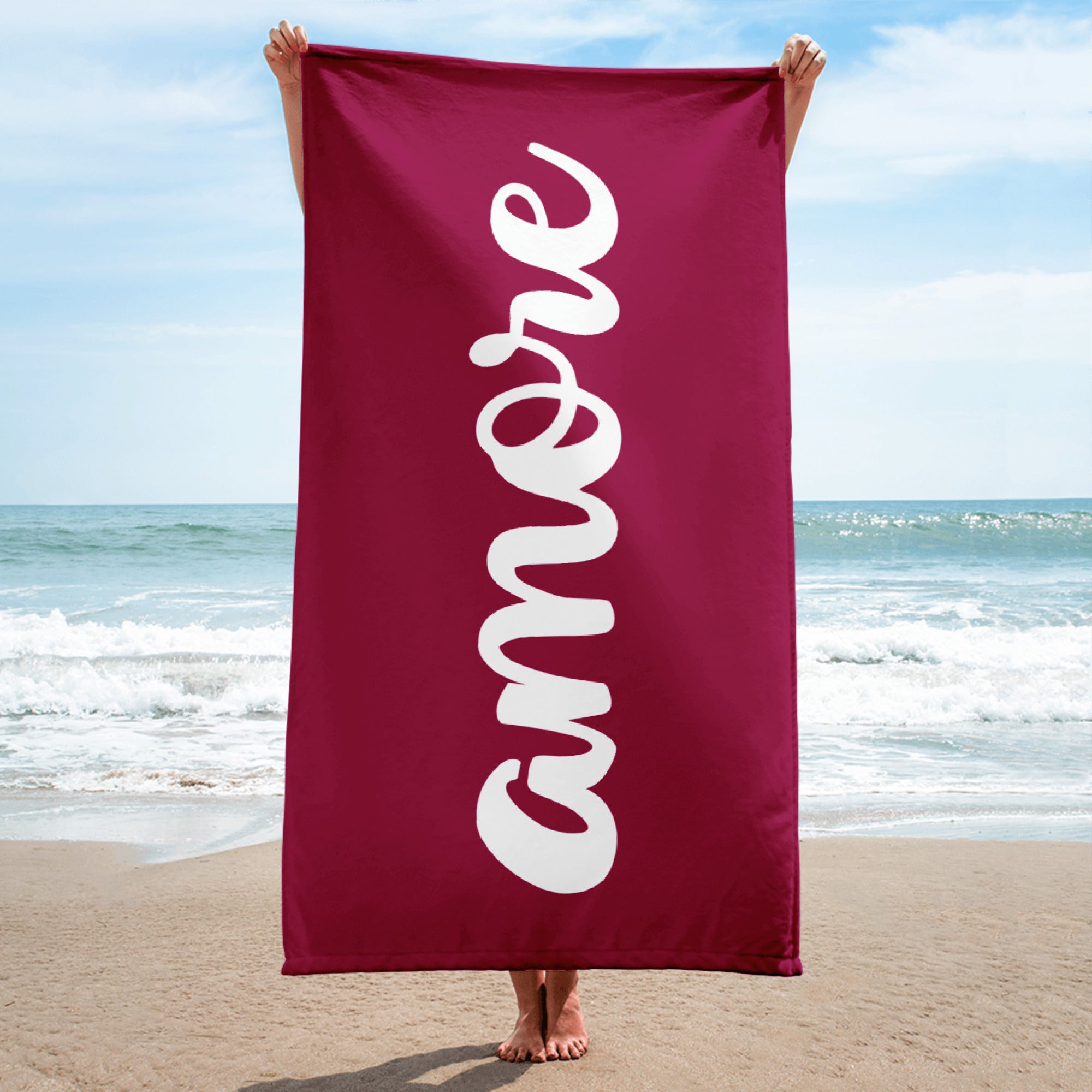 Person hält bordeauxfarbenes Handtuch mit weißem „Amore“-Schriftzug am Strand – Liebesbotschaft trifft italienisches Design.