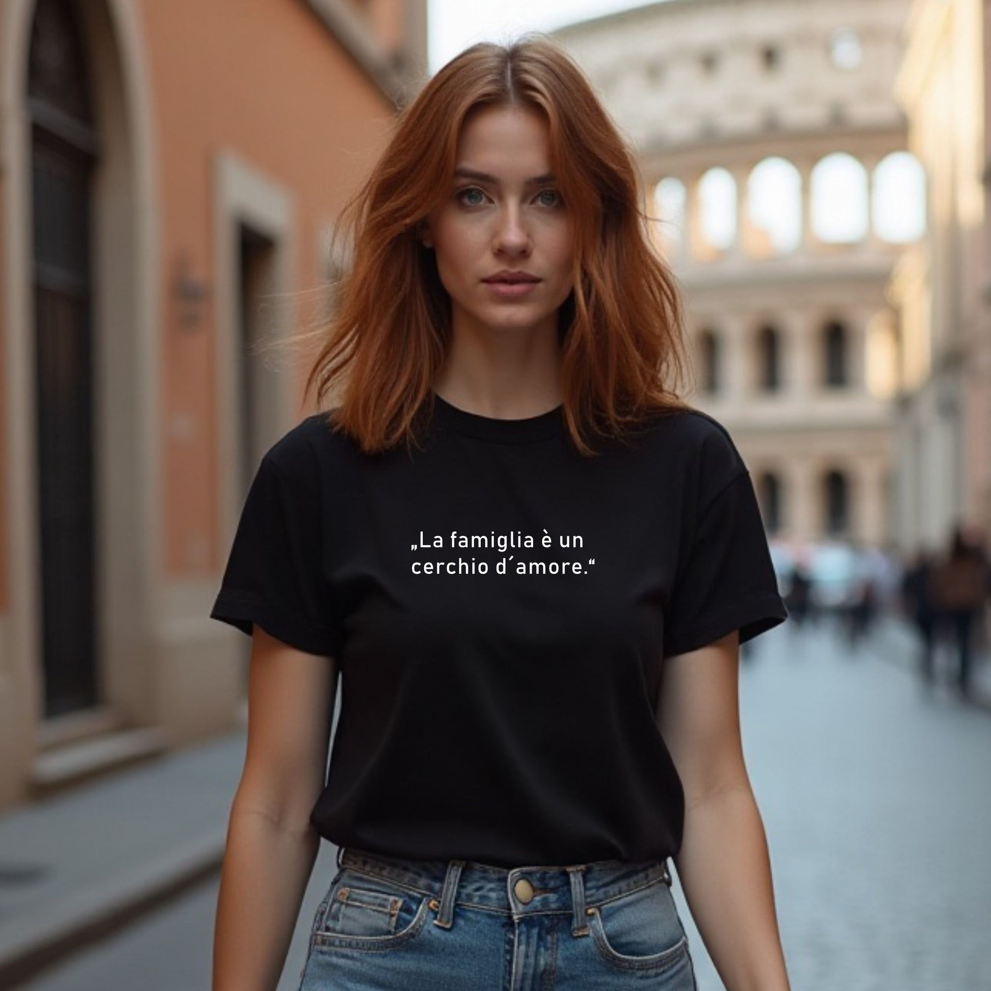Person mit schulterlangem Haar trägt schwarzes T-Shirt mit weißem italienischen Zitat „La famiglia è un cerchio d’amore“, fotografiert auf einer Kopfsteinpflasterstraße mit dem Kolosseum im Hintergrund